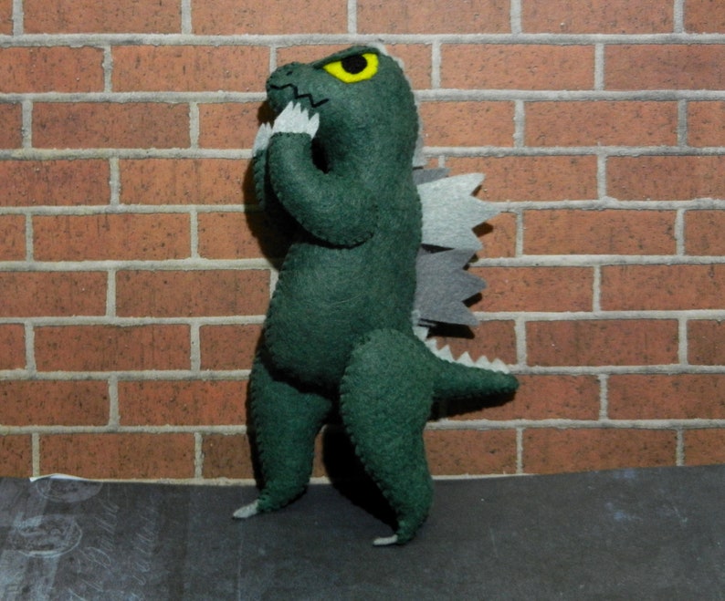 Puede incluir: Juguete monstruo de fieltro verde con un ojo amarillo y una espalda gris puntiaguda. El monstruo est&aacute; de pie sobre una superficie gris frente a una pared de ladrillo.