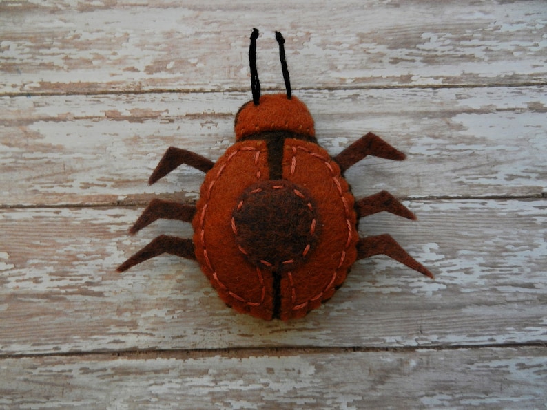 May include: A felt ladybug toy with brown and orange felt, and black felt antennae. The ladybug is sitting on a white wooden surface.