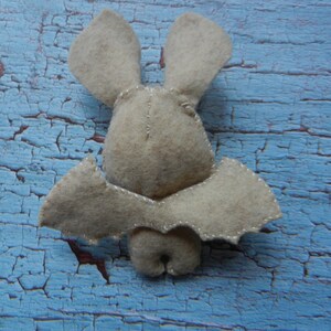 May include: A beige felt bat with stitched wings and ears. The bat is sitting on a blue, cracked, wooden surface.