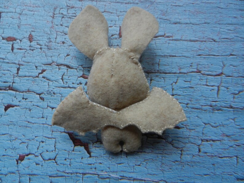 May include: A beige felt bat with stitched wings and ears. The bat is sitting on a blue, cracked, wooden surface.