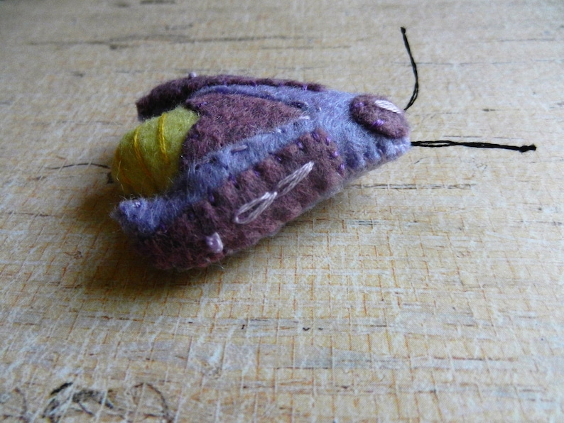 May include: A purple and yellow felt moth with black thread antennae.