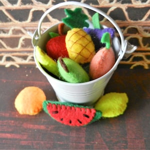 May include: A white bucket filled with felt fruit, including a pineapple, pear, apple, banana, watermelon, and two other fruits. The bucket is sitting on a brown wooden surface.