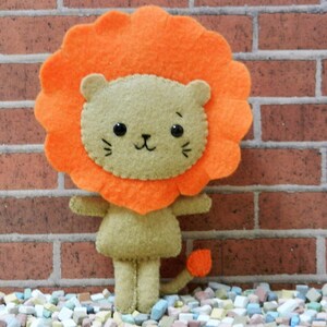 Può includere: Un leone di peluche con corpo marrone e criniera arancione. Il leone ha un viso carino e in stile cartone animato con occhi neri e un sorriso cucito.