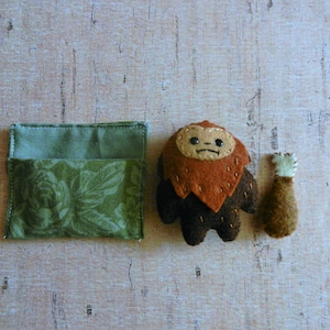 May include: A felt toy set featuring a brown and orange creature with a white face and a brown felt tail. The creature is standing next to a green fabric pouch with a floral pattern.