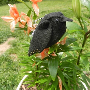 May include: A black felt bird with gold bead accents perched on a green plant with orange flowers.