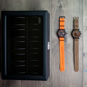 K&ouml;nnte beinhalten: Zwei Armbanduhren mit orangefarbenen und braunen B&auml;ndern auf einer Holzoberfl&auml;che. Die Uhren haben schwarze Zifferbl&auml;tter und silberne Zeiger. Der Text "modern gentleman" und "caddy bay collection" ist auf der Oberfl&auml;che sichtbar.