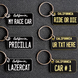 May include: Six black keychains with different text on them. The keychains are all in the shape of a license plate. The text on the keychains reads: "California My Race Car", "California Ride or Die", "California Pricilla", "California UR TXT HERE", "California LAZERCAT", and "California CAR # 1".