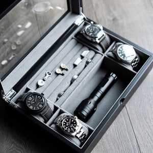 May include: A black wooden watch box with a glass top, lined with gray felt. The box contains four watches, a flashlight, and other small items.