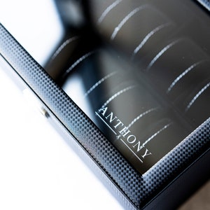 K&ouml;nnte beinhalten: Schwarze Uhrenbox mit durchsichtigem Glasdeckel und einem Karbonfaser-Muster. Die Box tr&auml;gt den Schriftzug "ANTHONY" auf der Vorderseite.