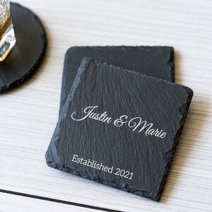 K&ouml;nnte beinhalten: Zwei schwarze Schieferuntersetzer mit dem Text "Justin & Marie Established 2021" in wei&szlig; graviert.