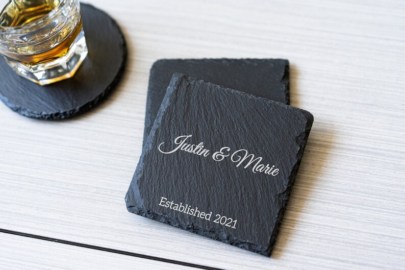 K&ouml;nnte beinhalten: Zwei schwarze Schieferuntersetzer mit dem Text "Justin & Marie Established 2021" in wei&szlig; graviert.
