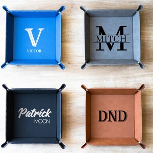 May include: Four personalized leather trays with different designs. The top left tray is blue with a white "V" and the name "VICTOR". The top right tray is gray with a white "M" and the name "MITCH". The bottom left tray is dark blue with a white "Patrick Moon" design. The bottom right tray is brown with a black "DND" design.