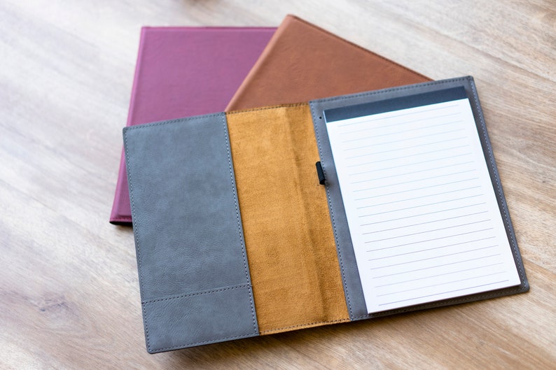 Op de afbeelding: Een grijs lederen notitieboek met een bruine leren flap en een wit gelinieerd notitieblok erin. Het notitieboek is open en het notitieblok is zichtbaar. Het notitieboek ligt op een houten oppervlak.