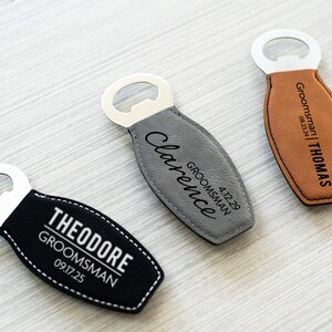 May include: Three personalised bottle openers with stainless steel tops and leather-like bottoms. One is black with white stitching and the name "THEODORE" and the text "GROOMSMAN 09.17.25". Another is grey with the name "Clarence" and the text "GROOMSMAN 4.12.29". The third is brown with the name "THOMAS" and the text "GROOMSMAN 08.23.24".