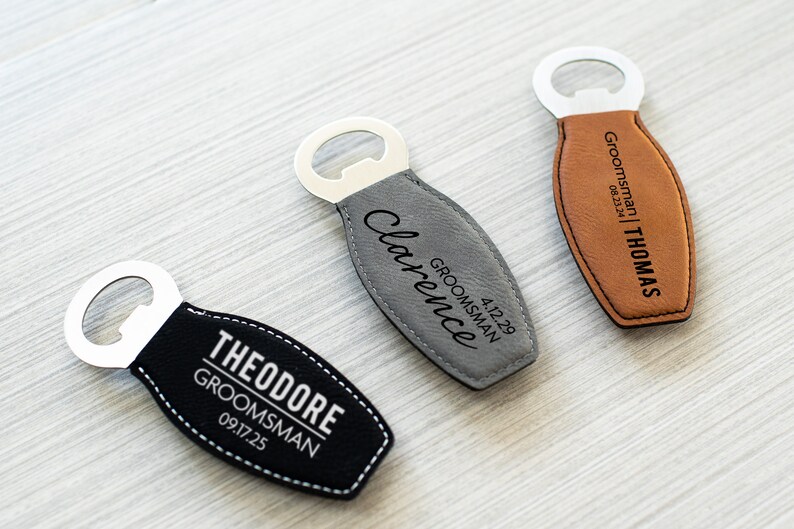 May include: Three personalised bottle openers with stainless steel tops and leather-like bottoms. One is black with white stitching and the name "THEODORE" and the text "GROOMSMAN 09.17.25". Another is grey with the name "Clarence" and the text "GROOMSMAN 4.12.29". The third is brown with the name "THOMAS" and the text "GROOMSMAN 08.23.24".