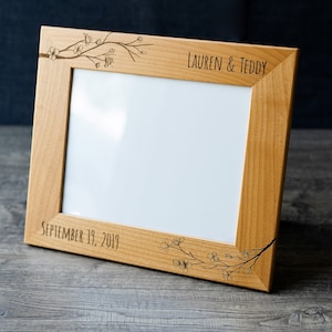 May include: A wooden picture frame with a white mat and a floral design engraved on the top and bottom. The frame is personalized with the text "LAUREN & TEDDY" and "SEPTEMBER 19, 2019".