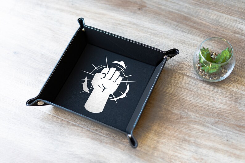May include: Black leather dice tray with a white fist graphic and white stitching around the edges. The tray is on a light wood surface.