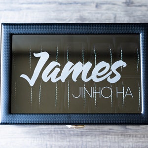May include: A black rectangular box with a mirrored lid. The lid has the name "James" written in white cursive lettering and the name "JINHO HA" written in white block lettering below it.