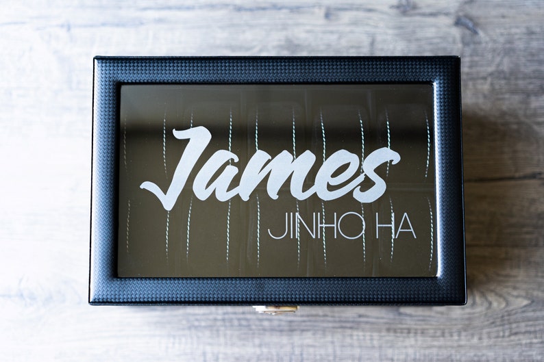May include: A black rectangular box with a mirrored lid. The lid has the name "James" written in white cursive lettering and the name "JINHO HA" written in white block lettering below it.