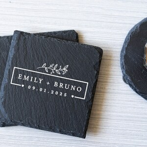 K&ouml;nnte beinhalten: Zwei schwarze Schieferuntersetzer mit einem wei&szlig;en Design. Das Design beinhaltet einen Blumenzweig, den Text "Emily + Bruno" und das Datum "09.01.2025".
