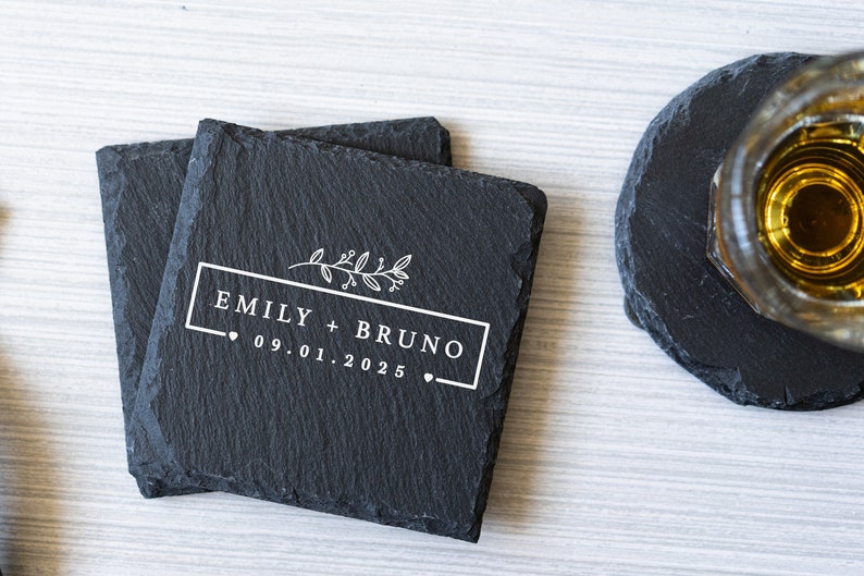 K&ouml;nnte beinhalten: Zwei schwarze Schieferuntersetzer mit einem wei&szlig;en Design. Das Design beinhaltet einen Blumenzweig, den Text "Emily + Bruno" und das Datum "09.01.2025".