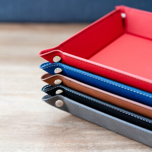 May include: A stack of four leather trays in different colors, including red, blue, brown, and gray. Each tray has a stitched edge and metal rivets.
