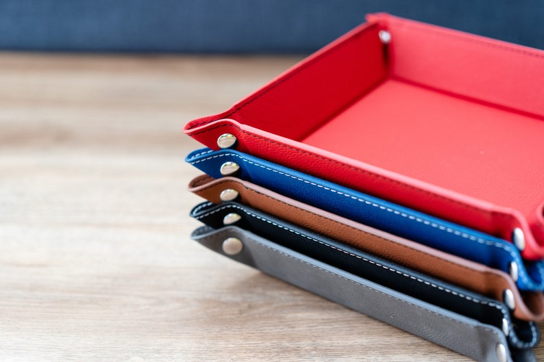 May include: A stack of four leather trays in different colors, including red, blue, brown, and gray. Each tray has a stitched edge and metal rivets.