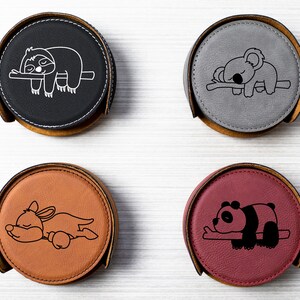 May include: Set of four round leather coasters with different animal designs. The coasters are black, gray, brown, and burgundy. Each coaster features a different animal sleeping on a branch, including a sloth, koala, kangaroo, and panda.