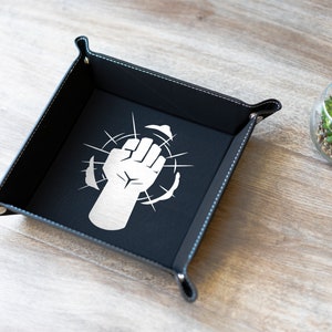 May include: Black leather dice tray with a white fist graphic and white stitching around the edges. The tray is on a light wood surface.