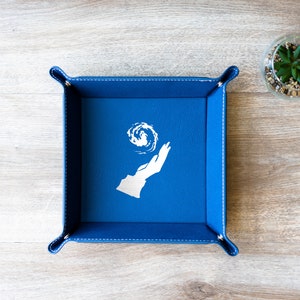 May include: A blue felt dice tray with a white design of a hand holding a swirling orb. The tray has a stitched border.
