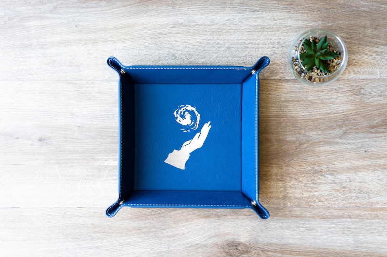 May include: A blue felt dice tray with a white design of a hand holding a swirling orb. The tray has a stitched border.