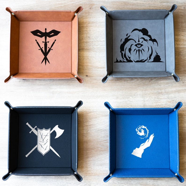 May include: Four different colored felt dice trays with various designs. The trays are square and have a raised edge. The designs include a mask with swords, an eye with a cloud, a shield with a sword and axe, and a hand with a swirling symbol.