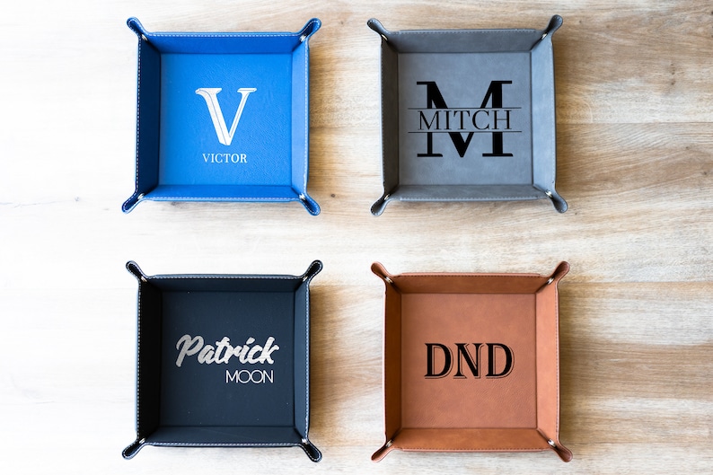 May include: Four personalized leather trays with different designs. The top left tray is blue with a white "V" and the name "VICTOR". The top right tray is gray with a white "M" and the name "MITCH". The bottom left tray is dark blue with a white "Patrick Moon" design. The bottom right tray is brown with a black "DND" design.