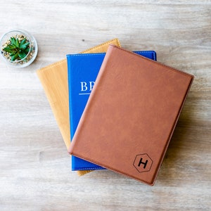 Op de afbeelding: Drie lederen notitieboekjes gestapeld op elkaar. Het bovenste notitieboekje is bruin met een zeshoekig monogram van de letter "H" gegraveerd op de voorzijde. Het middelste notitieboekje is blauw met het woord "BRAND" in wit gedrukt op de voorzijde. Het onderste notitieboekje is bruin met een houtnerfpatroon.