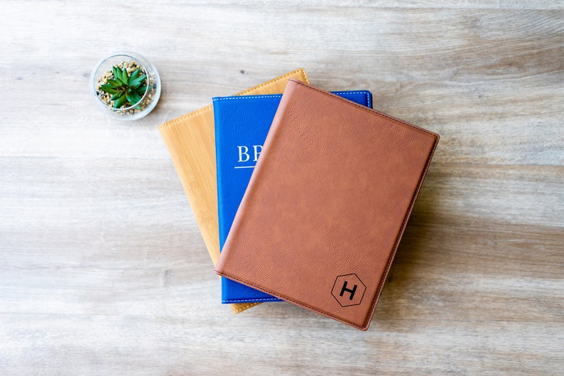Op de afbeelding: Drie lederen notitieboekjes gestapeld op elkaar. Het bovenste notitieboekje is bruin met een zeshoekig monogram van de letter "H" gegraveerd op de voorzijde. Het middelste notitieboekje is blauw met het woord "BRAND" in wit gedrukt op de voorzijde. Het onderste notitieboekje is bruin met een houtnerfpatroon.
