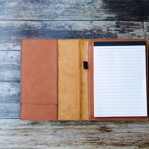 Op de afbeelding: Een bruin lederen gebonden notitieboek met een blanco, gestreepte pagina open. Het notitieboek ligt op een houten oppervlak.