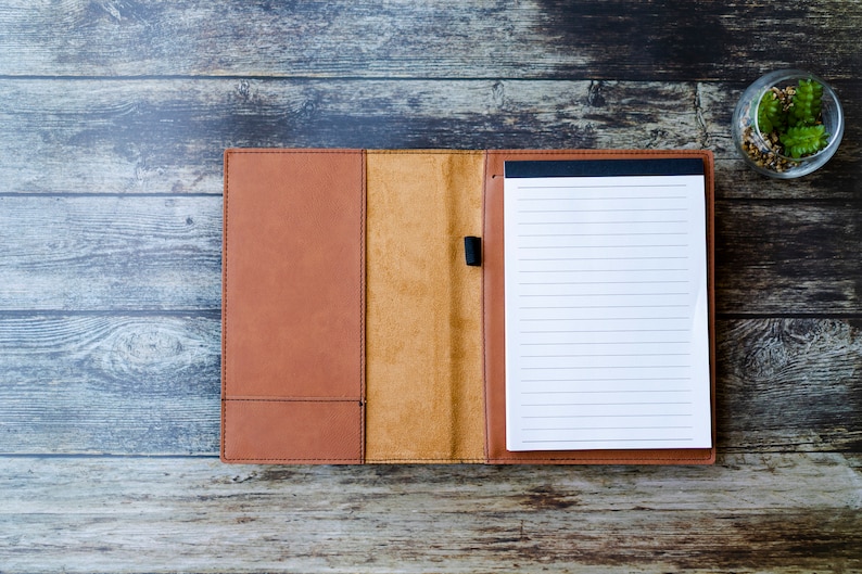 Op de afbeelding: Een bruin lederen gebonden notitieboek met een blanco, gestreepte pagina open. Het notitieboek ligt op een houten oppervlak.