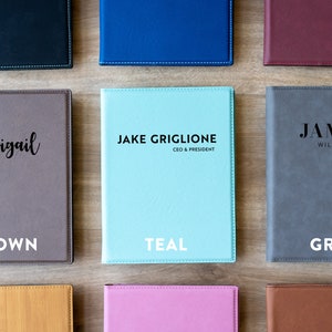 Op de afbeelding: Negen verschillende gekleurde kunstlederen notitieboekjes met verschillende namen en titels op de voorkant. De kleuren omvatten bruin, teal, grijs, oranje, roze en bruin.
