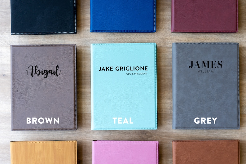 Op de afbeelding: Negen verschillende gekleurde kunstlederen notitieboekjes met verschillende namen en titels op de voorkant. De kleuren omvatten bruin, teal, grijs, oranje, roze en bruin.
