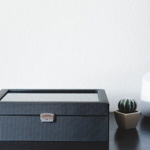 K&ouml;nnte beinhalten: Eine schwarze Uhrenbox mit Glasdeckel und silbernem Verschluss. Die Box steht auf einer dunklen Holzoberfl&auml;che neben einem wei&szlig;en intelligenten Lautsprecher und einem kleinen Topf mit Kaktus.