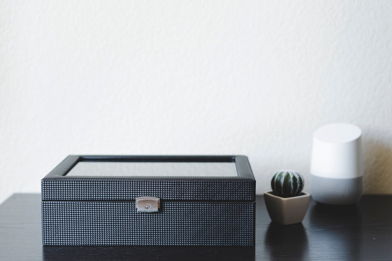K&ouml;nnte beinhalten: Eine schwarze Uhrenbox mit Glasdeckel und silbernem Verschluss. Die Box steht auf einer dunklen Holzoberfl&auml;che neben einem wei&szlig;en intelligenten Lautsprecher und einem kleinen Topf mit Kaktus.