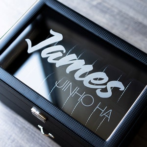 May include: A black watch box with a clear glass top. The box has a carbon fiber pattern and the name "James JinHo Ha" is engraved on the glass.