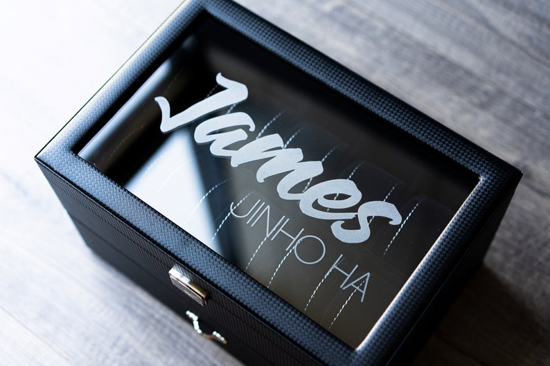 May include: A black watch box with a clear glass top. The box has a carbon fiber pattern and the name "James JinHo Ha" is engraved on the glass.