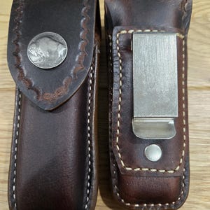 May include: Two brown leather knife sheaths. One has a silver buffalo nickel and decorative stitching. The other has a silver metal clip and white stitching. Both are closed and appear to be new.