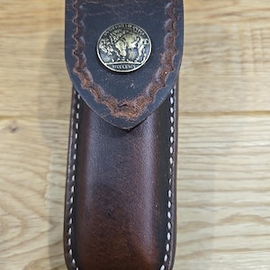 May include: A brown leather knife sheath with white stitching and a metal button featuring an image of a buffalo with the text "United States of America" and "Five Cents".