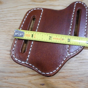 May include: Brown leather knife sheath with white stitching. The sheath is measured with a tape measure, showing it is 2 inches wide.