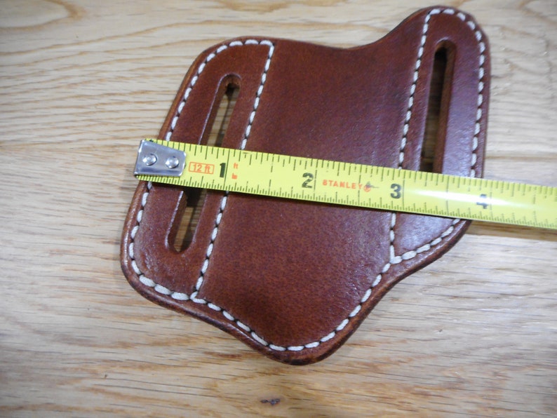 May include: Brown leather knife sheath with white stitching. The sheath is measured with a tape measure, showing it is 2 inches wide.