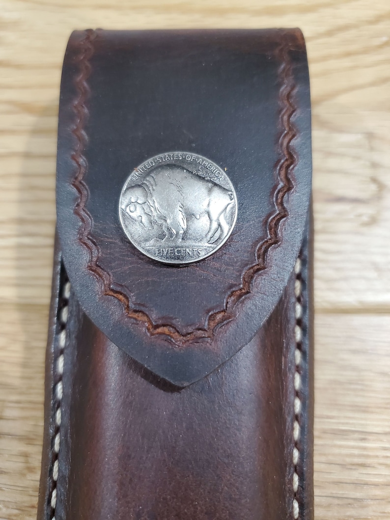 Peut inclure: Un &eacute;tui en cuir marron avec des d&eacute;tails de couture blancs. Une pi&egrave;ce de monnaie en nickel de buffle argent&eacute;e est fix&eacute;e &agrave; l'avant de l'&eacute;tui.