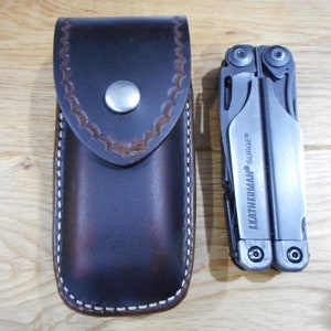 May include: A brown leather sheath with white stitching and a snap closure. The sheath is designed to hold a multi-tool. A silver multi-tool with the text "LEATHERMAN SURGE" is shown next to the sheath.