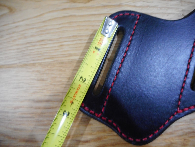 May include: A black leather holster with red stitching and a belt loop. The holster is being measured with a tape measure, showing a measurement of 3 inches.